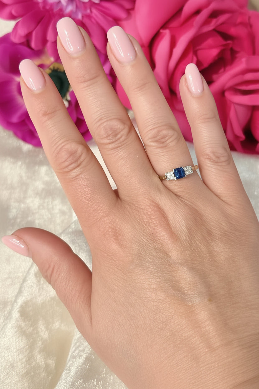 Hand wearing a sapphire and old european cut diamond trilogy ring in front of a pink flower