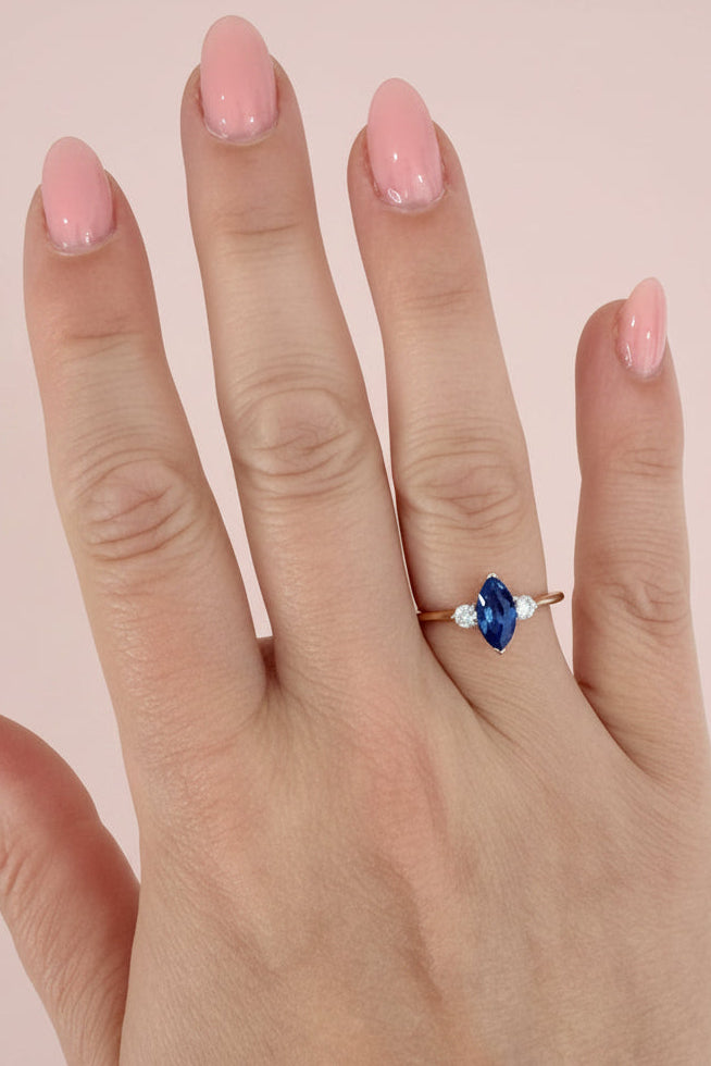 Hand wearing a ring with a blue sapphire on a textured pale pink background
