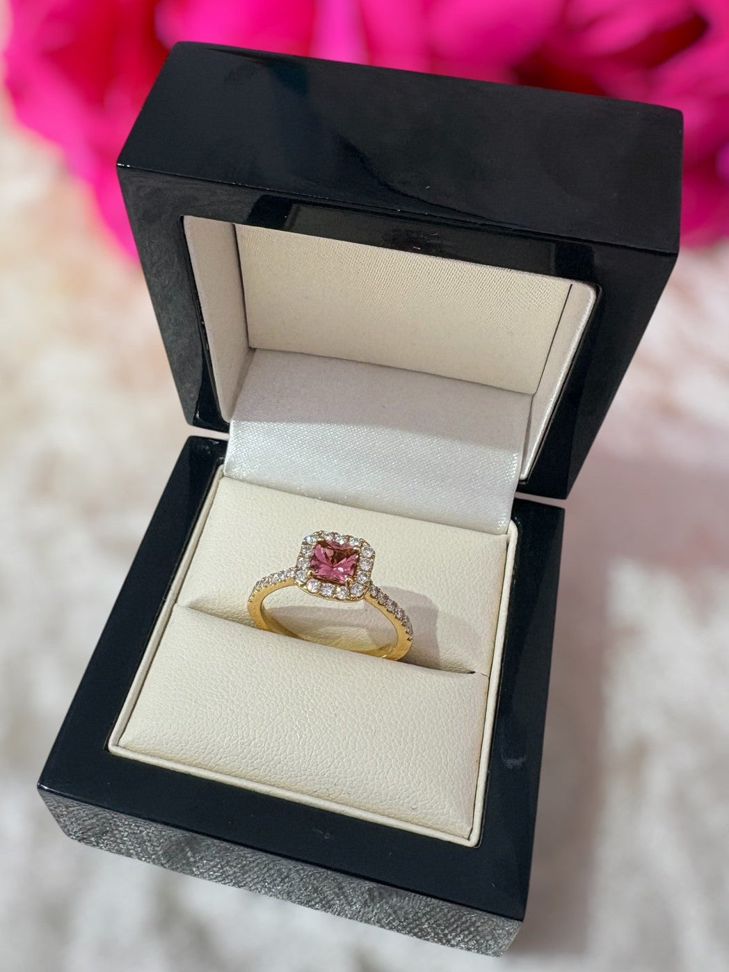 Gold ring with a pink gemstone in a black jewelry box on a soft surface with pink flowers in the background.