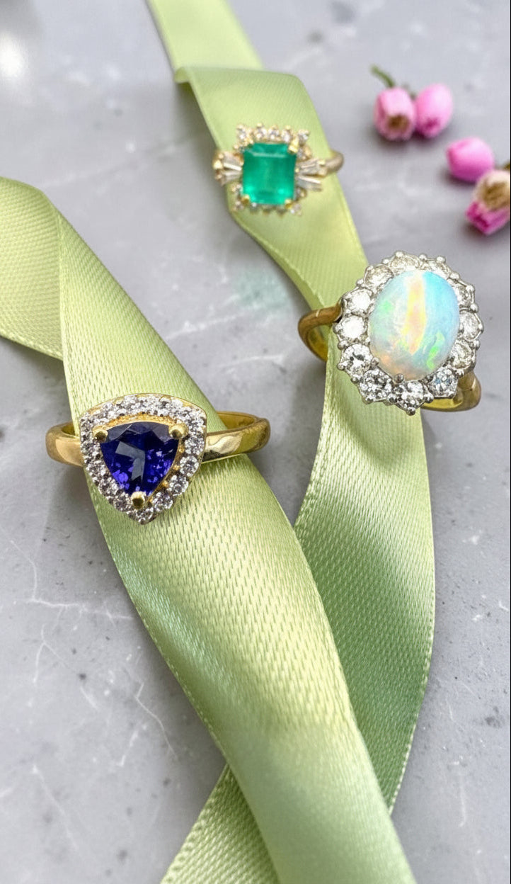Three gold rings with colorful gemstones are displayed on a light green satin ribbon against a gray marble background. The rings feature a heart-shaped blue sapphire, a square green emerald, and an oval opal surrounded by clear crystals.