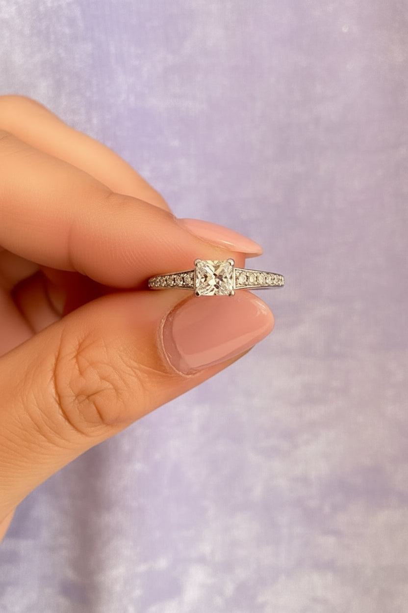 princess diamind ring with diamond shoulders being held between two fingers against a lilac background
