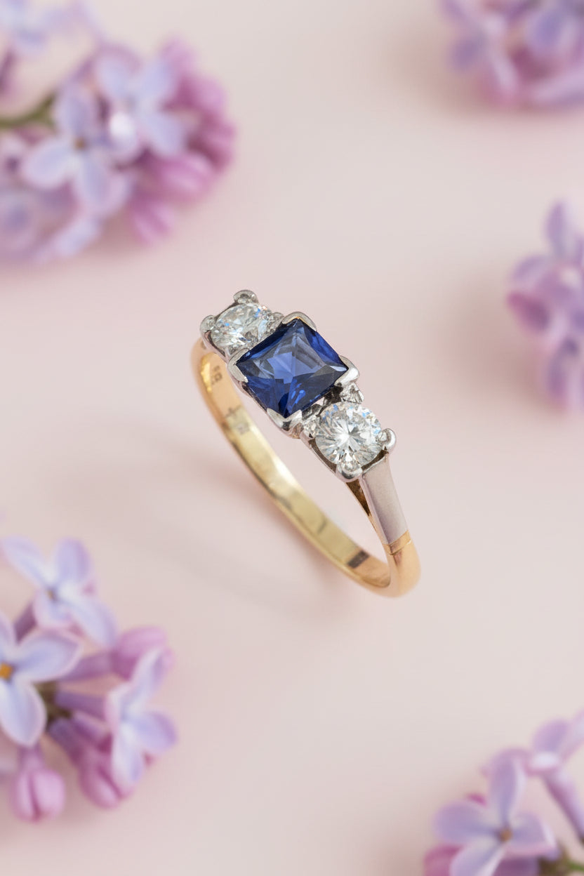 18ct Gold ring with a blue sapphire and old European cut diamonds on a pink background with lilace flowers 