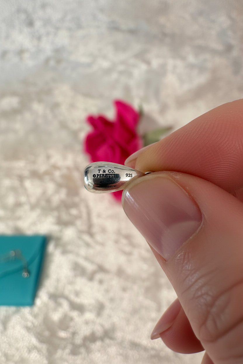 Close-up of a hand holding a clear teardrop-shaped pendant with a  hallmake 'T & Co' and '925' on a blurred background