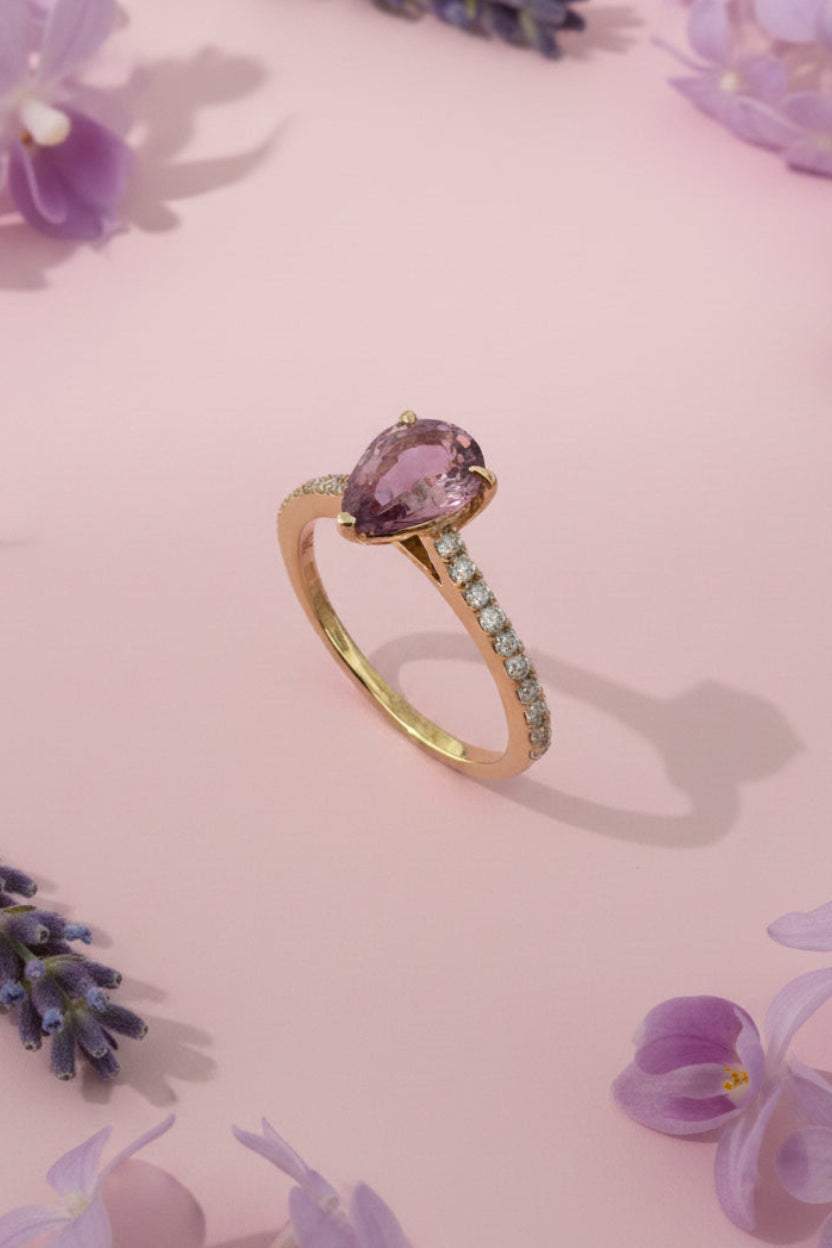Gold ring with brown sapphire and diamond shoulders 
on a pink surface with lilac flowers