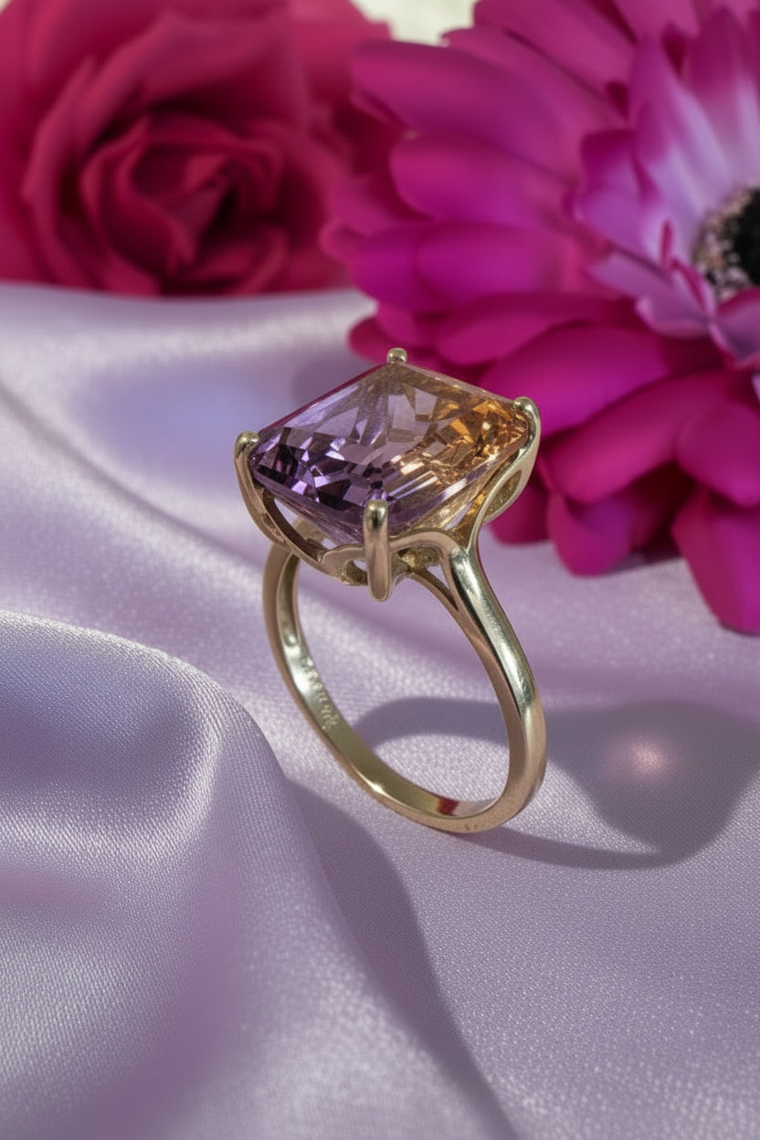 A gold ring with an emerald cut ametrine gemstone on a lilac silk surface with pink flowers