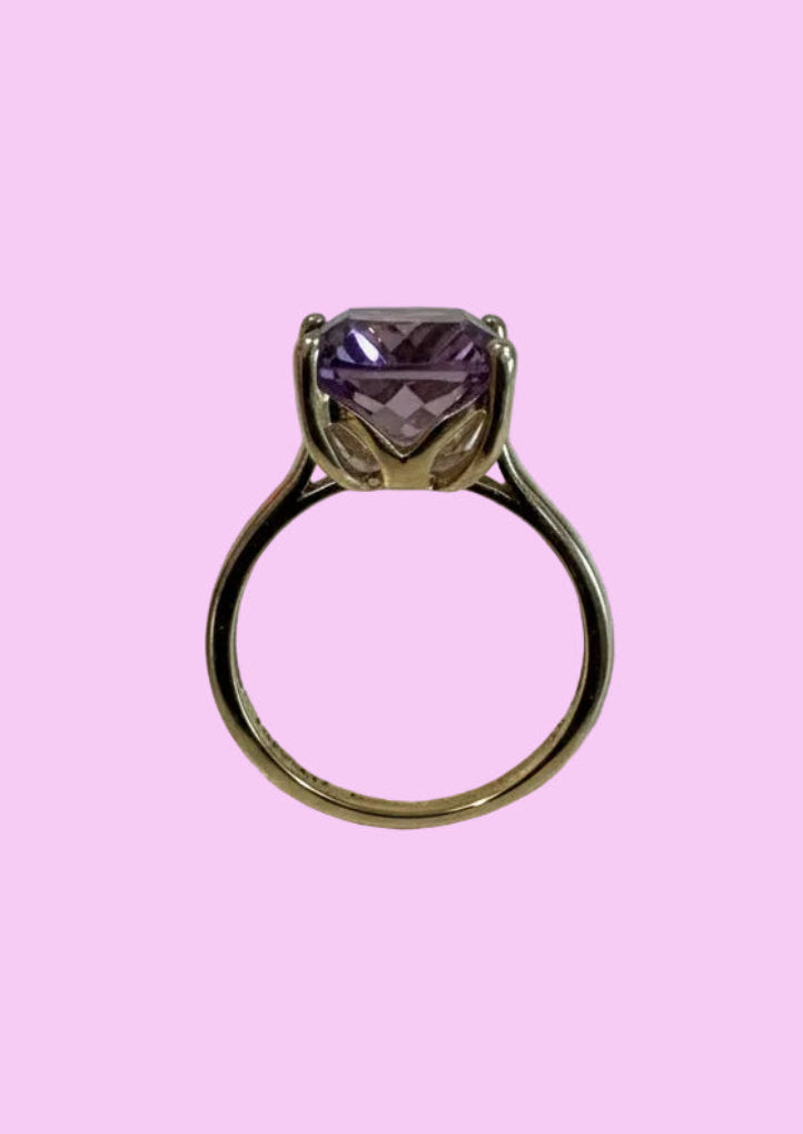 A gold ring with a large faceted ametrine gemstone, centered against a solid light pink background.