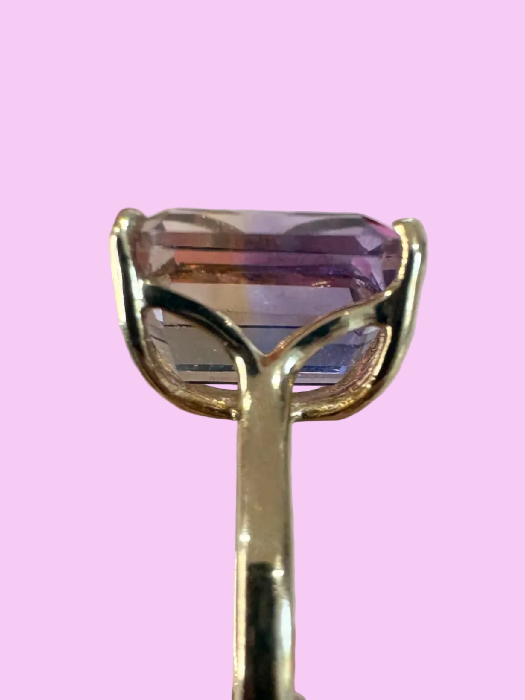 A gold ring with a large rectangular purple gemstone held by tweezers, with a hand holding the tweezers on the right side and pink flowers on a textured white background.