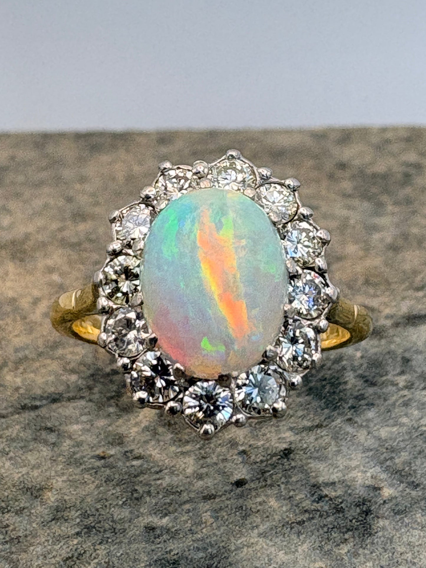 A vintage-style ring featuring an oval opal gemstone with iridescent rainbow play-of-colour, surrounded by a halo of diamonds and a gold band, shown on a textured grey stone surface.