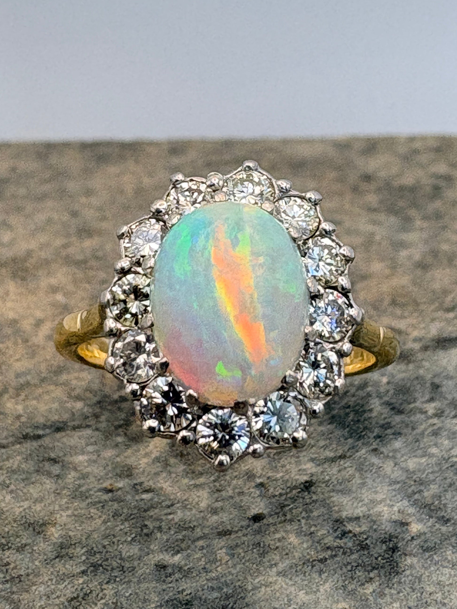 A vintage-style ring featuring an oval opal gemstone with iridescent rainbow play-of-colour, surrounded by a halo of diamonds and a gold band, shown on a textured grey stone surface.