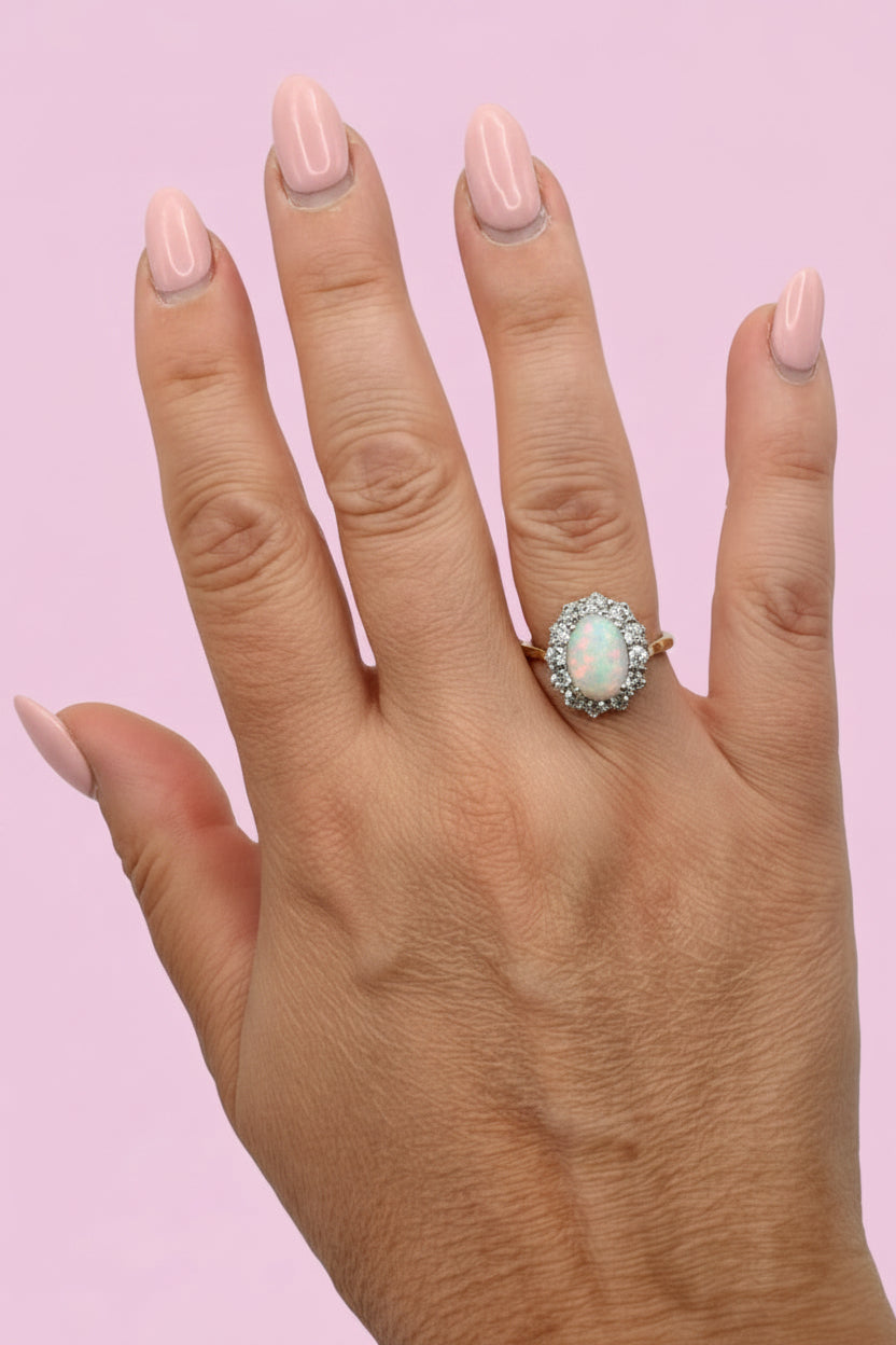 A close-up of a hand with mint green painted nails wearing a gold ring with a large opal stone surrounded by diamonds, set against a plain light pink background.