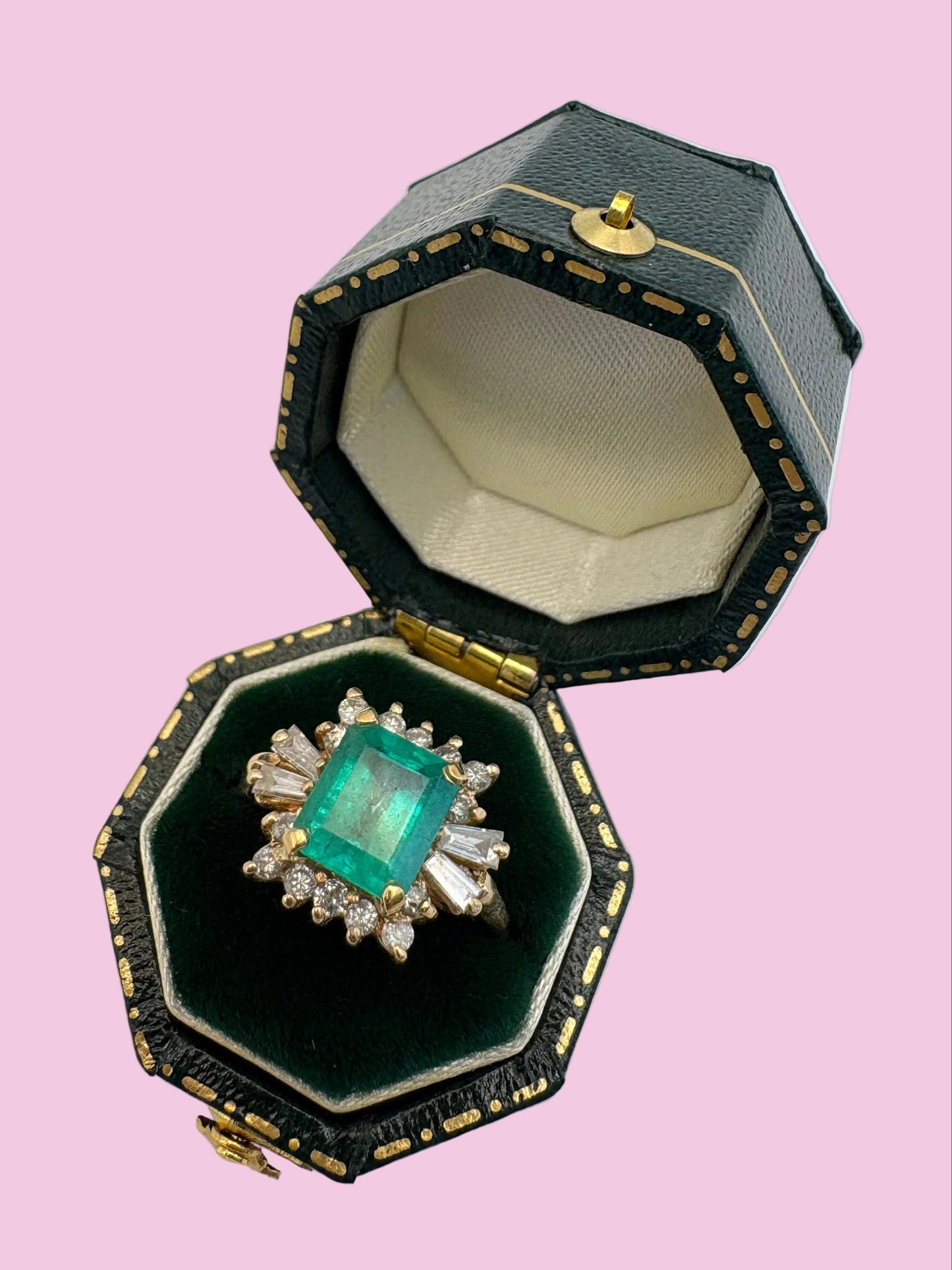 An open octagonal black jewelry box with gold trim and a green velvet interior, displaying a ring with a large green emerald-cut center stone and clear surrounding stones on a light gray background.