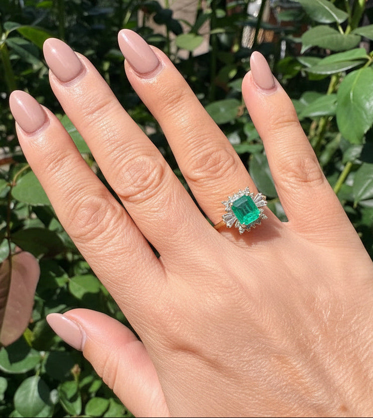 A silver gold ring with a rectangular green emerald-cut emerald and small diamonds, shown worn on a hand with mint greenneautral nail polish against a background of green foliage.