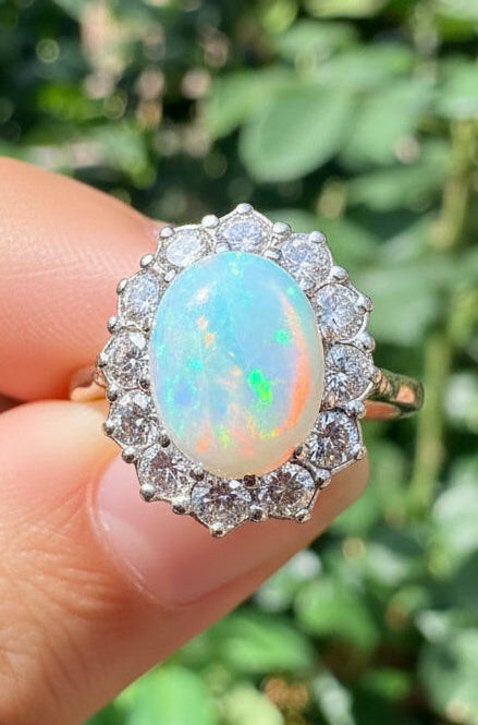A close-up of a hand holding a ring with a large iridescent opal center stone, set against a blurred background of green foliage.