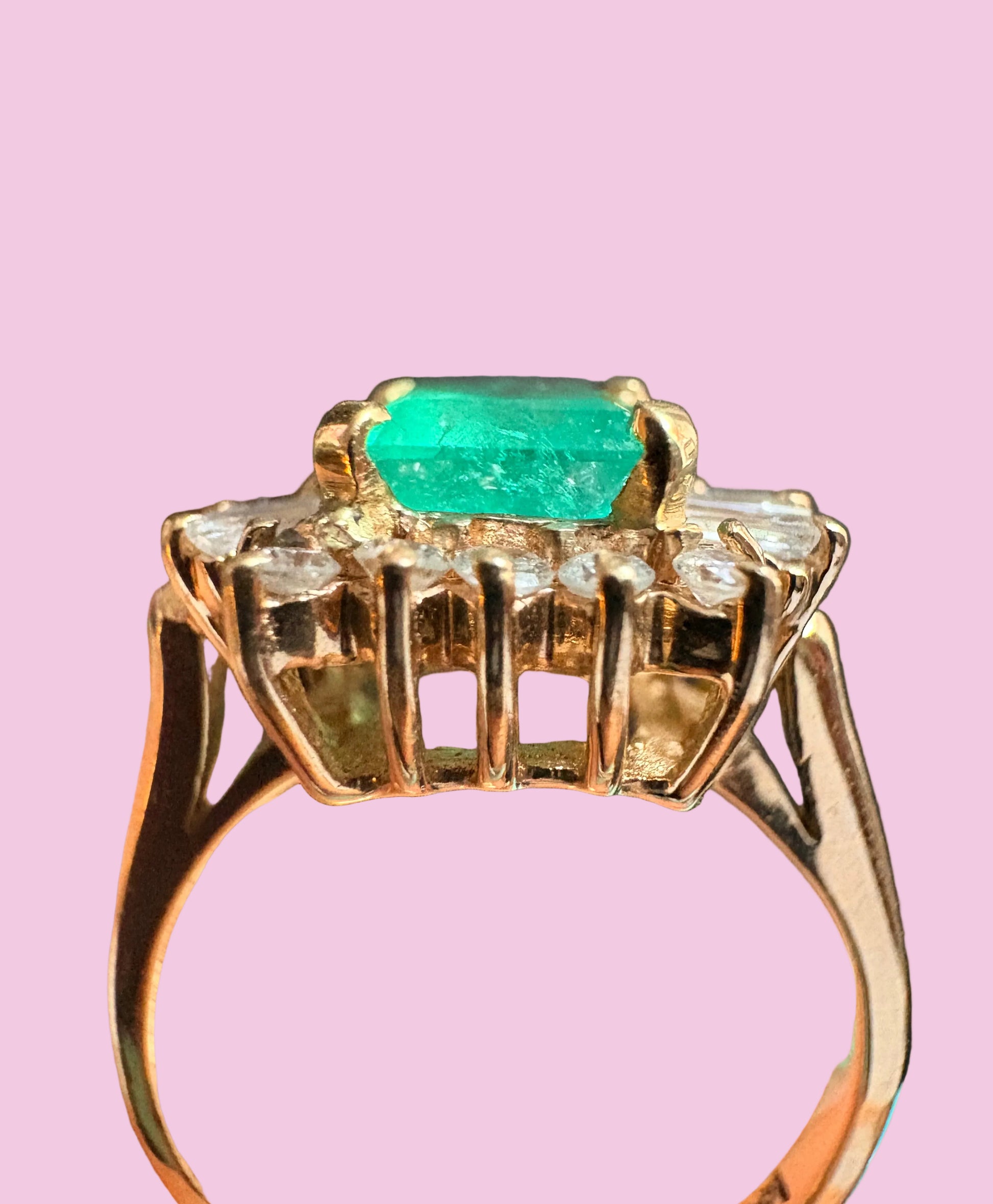 A yellow gold ring featuring a rectangular emerald-cut green gemstone in the center, flanked by small clear stones and a decorative openwork top, shown against a solid pink background.