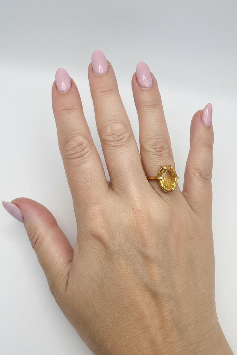 A close-up of a person's hand wearing a yellow gold ring with an oval-cut yellow gemstone, set against a plain light gray background.