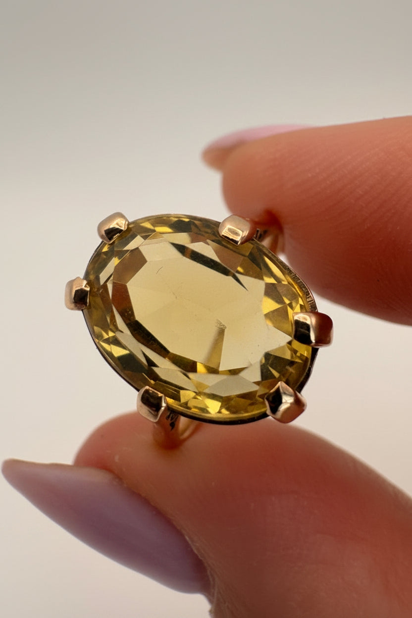A close-up of a yellow oval gemstone ring held between two fingers against a plain light background.