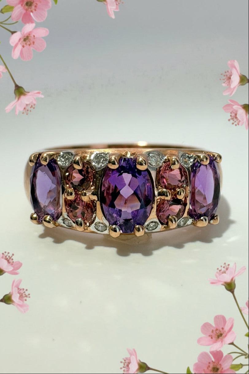 A rose gold ring featuring a row of oval amethyst and pink tournaline stones with small diamond accent stones, shown on a plain light background with pink flowers.