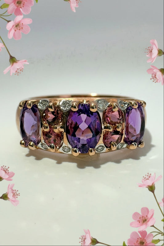 A rose gold ring featuring a row of oval amethyst and pink tournaline stones with small diamond accent stones, shown on a plain light background with pink flowers.