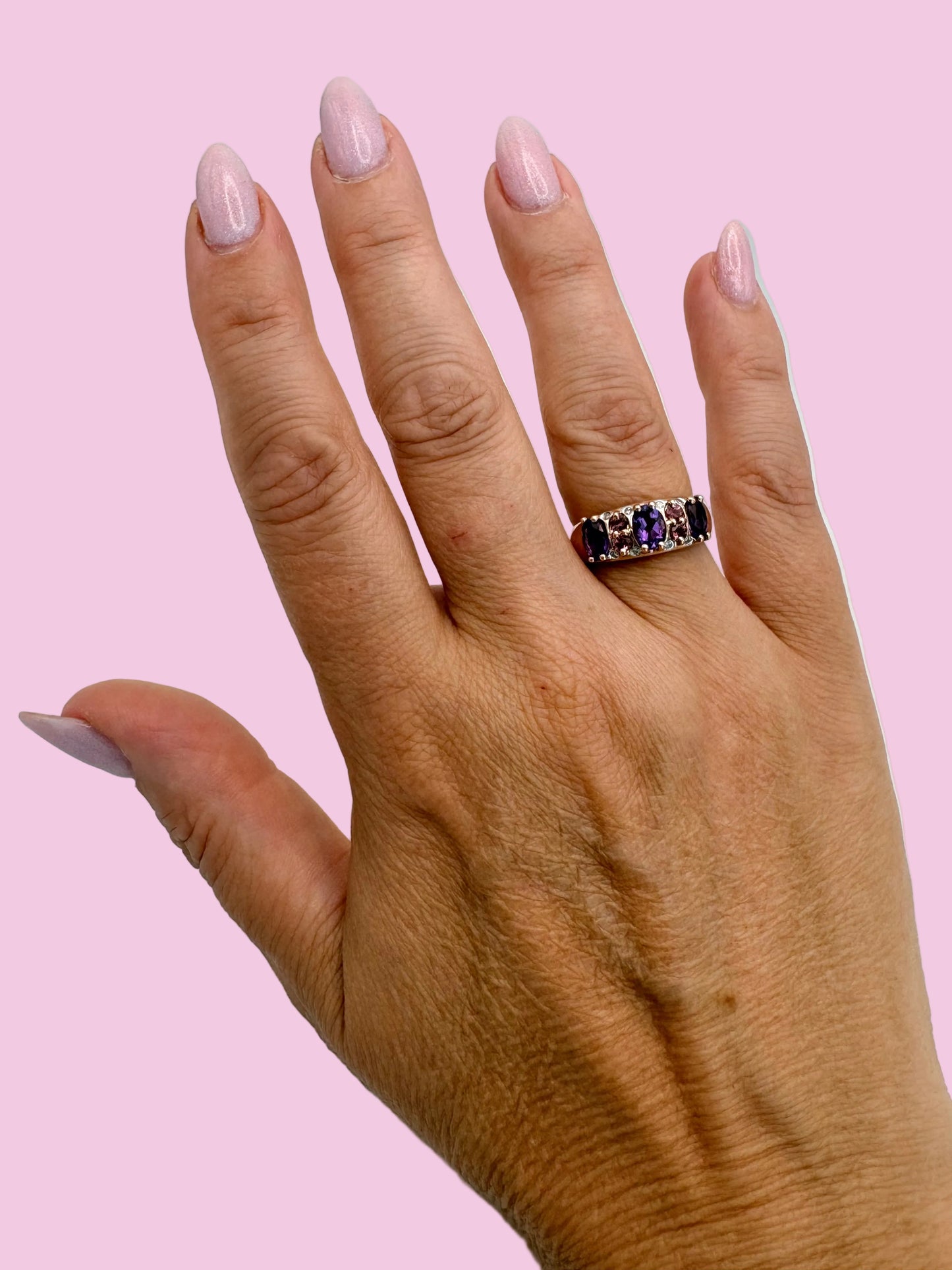 A close-up of a hand wearing a rose gold band ring set with amethyst, tourmaline and diamond stones, photographed against a light purple background.