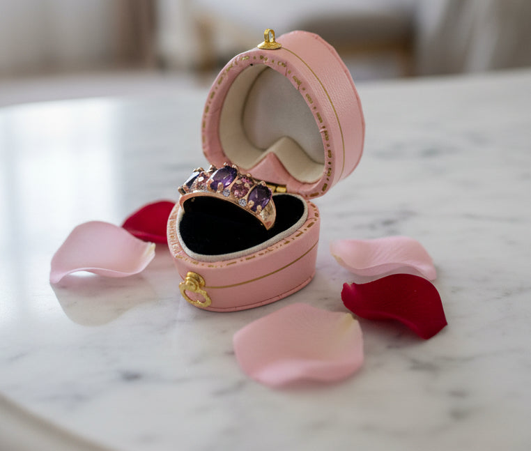 A pink heart-shaped ring box with gold trim and a black velvet interior, shown open on a white background. A gold ring with large purple and red gemstones is displayed inside the box.