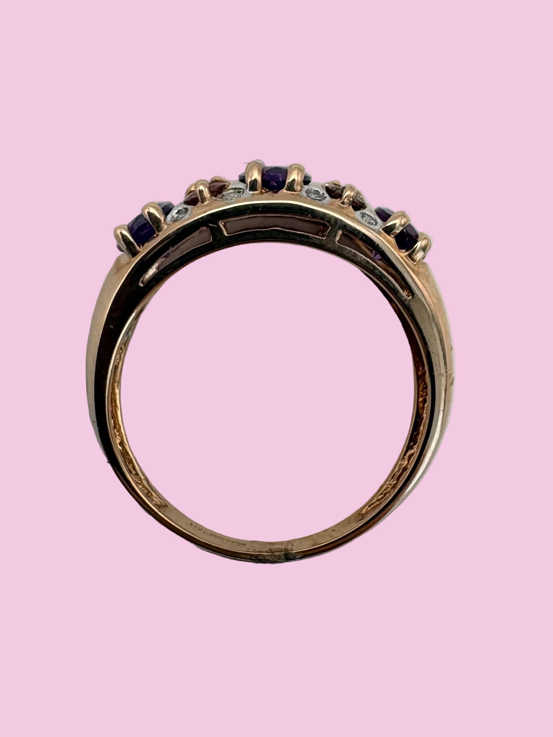 A rose gold ring with a row of amethyst, tourmaline and diamond accents shown from a top-down view on a light pink background, 