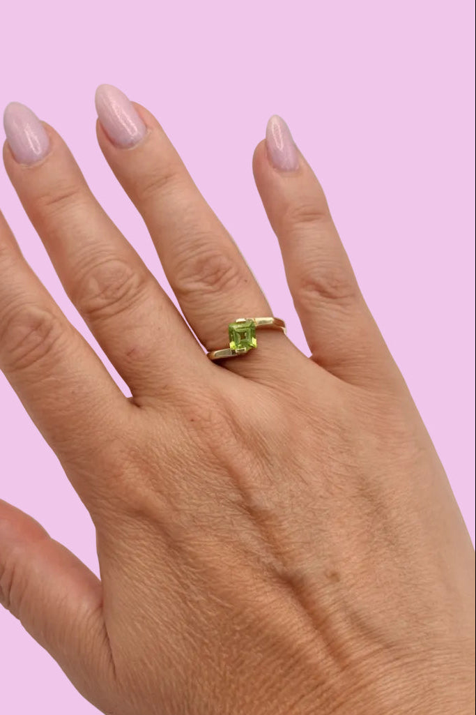 A close-up of a hand with pale pink manicured nails wearing a gold ring with a square-cut green gemstone, set against a solid light pink background.