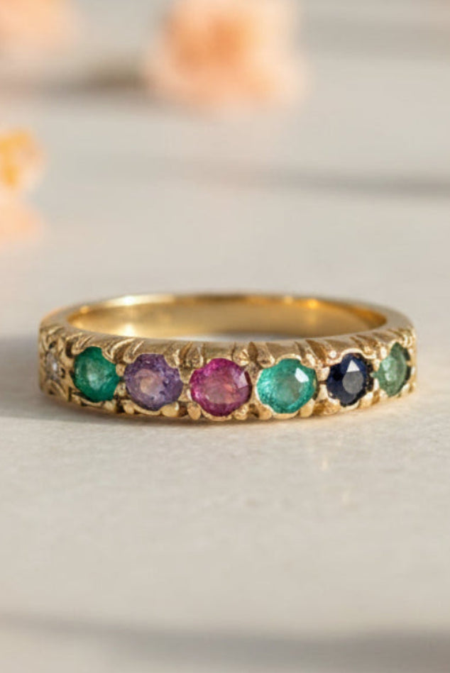 A gold band ring featuring a row of round gemstones in Diamond, Emerald, Amethyst, Ruby, Emerald, Sapphire and Tourmaline thereby spelling DEAREST. set against a soft-focus light grey background with blurred peach flowers.