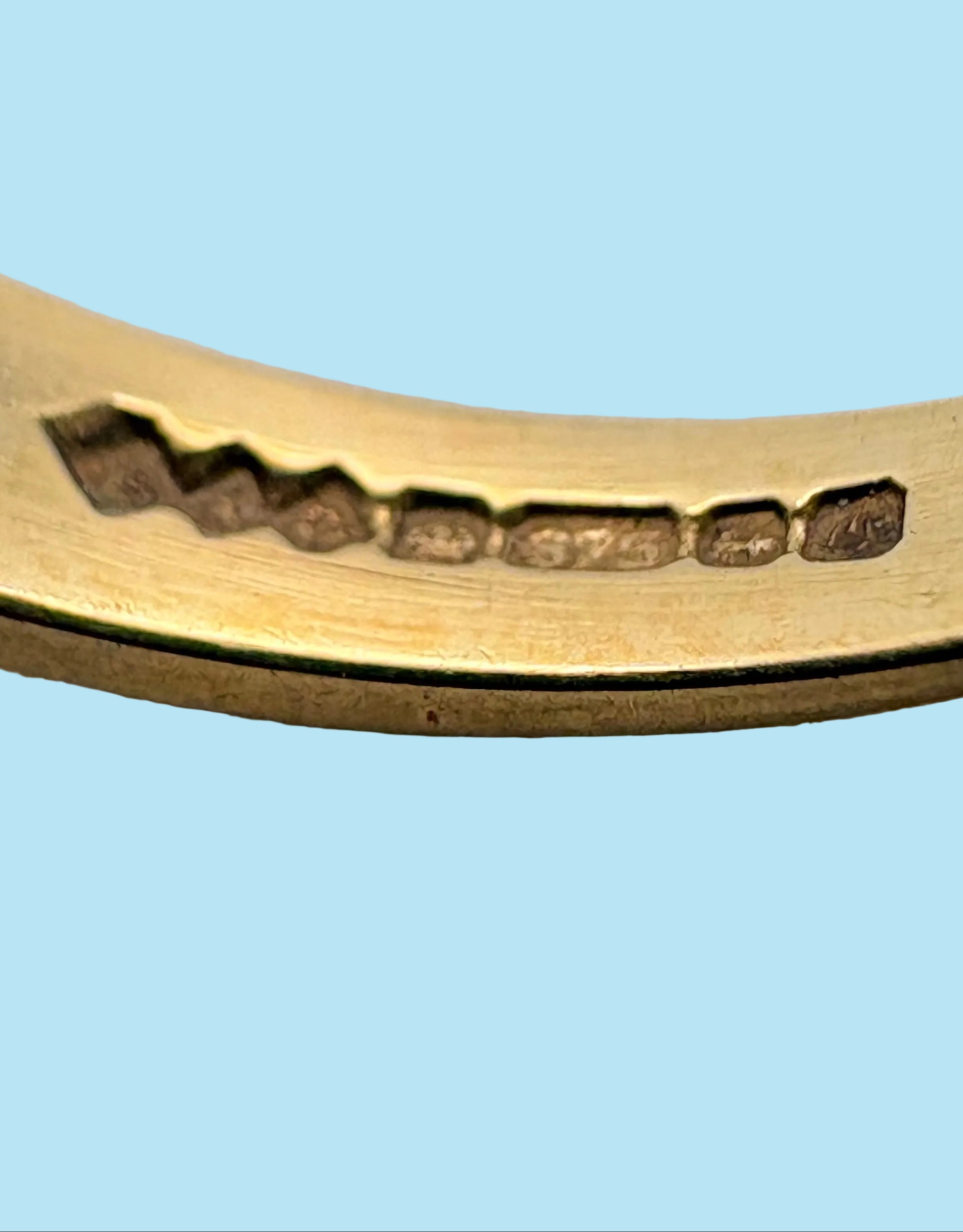 Close-up of a curved gold-toned metal band with a row of small dark rectangular inlays or markings along the inner side, photographed on a light gray background.