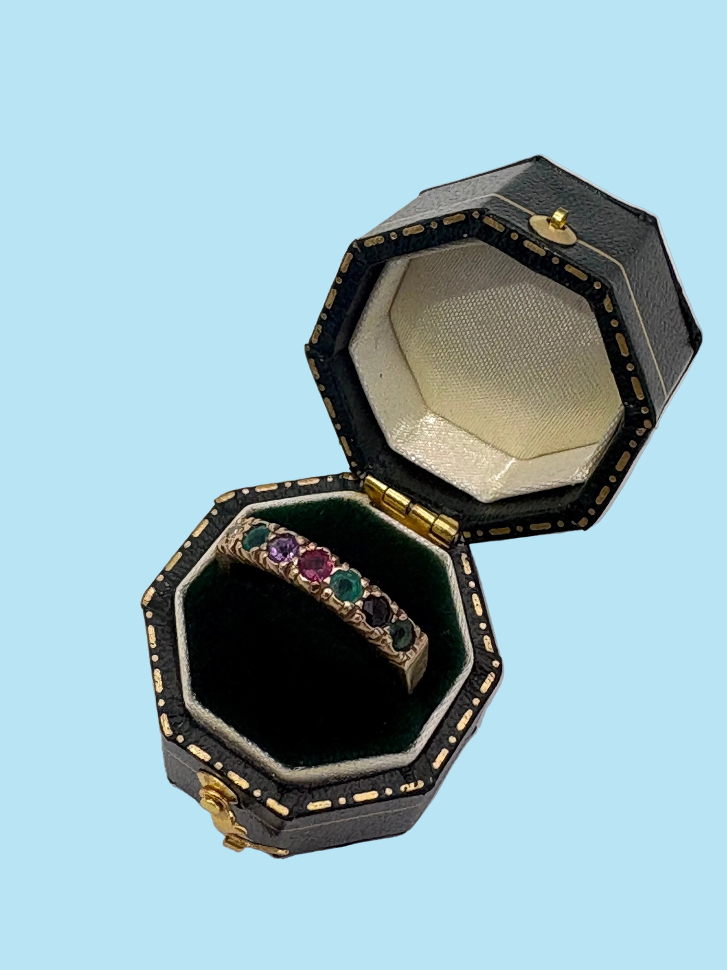 An open octagonal black jewelry box with gold trim and a cream velvet interior, displaying a gold ring set with multiple colored gemstones on a light gray background.