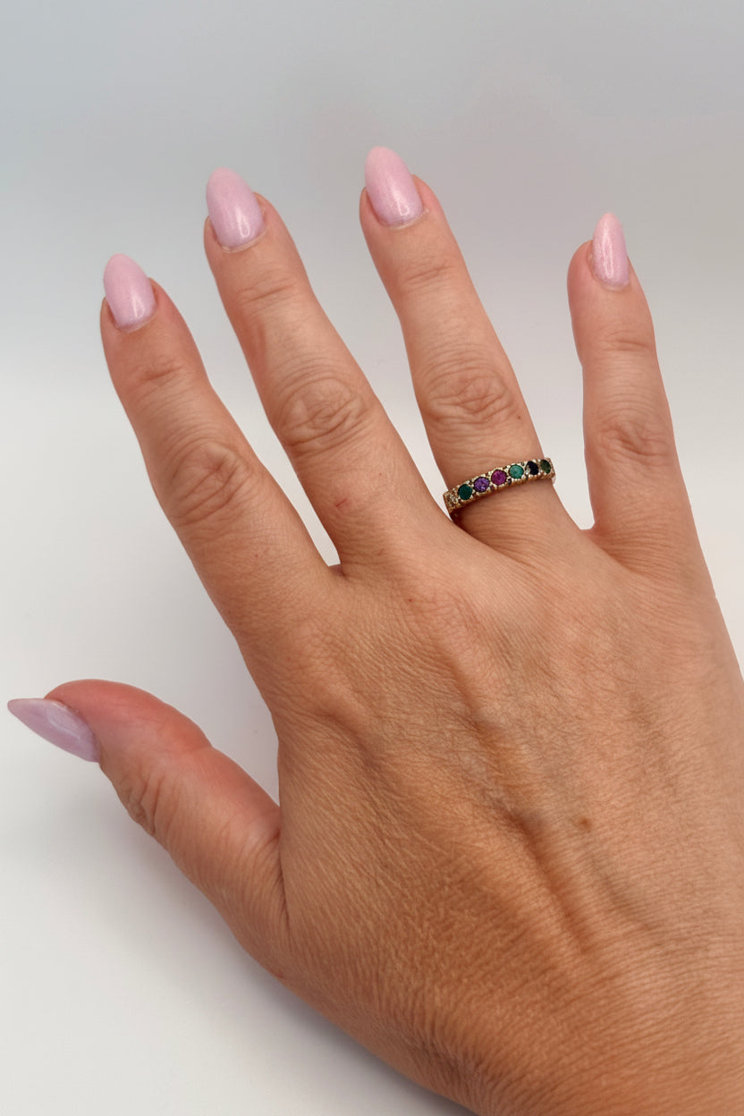 A close-up of a person's hand wearing a thin gold band ring set with small multicolored gemstones, shown against a plain light gray background.