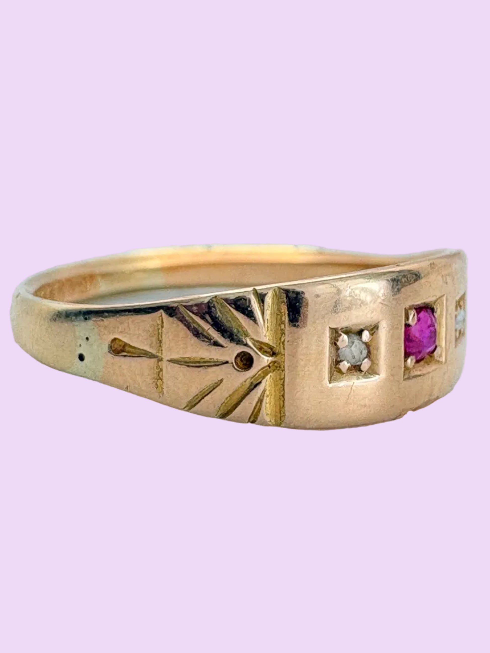 A antique yellow gold ring with engraved geometric and leaf motifs, featuring a small round clear stone and a square-cut pink gemstone on the band, set against a light blue background.