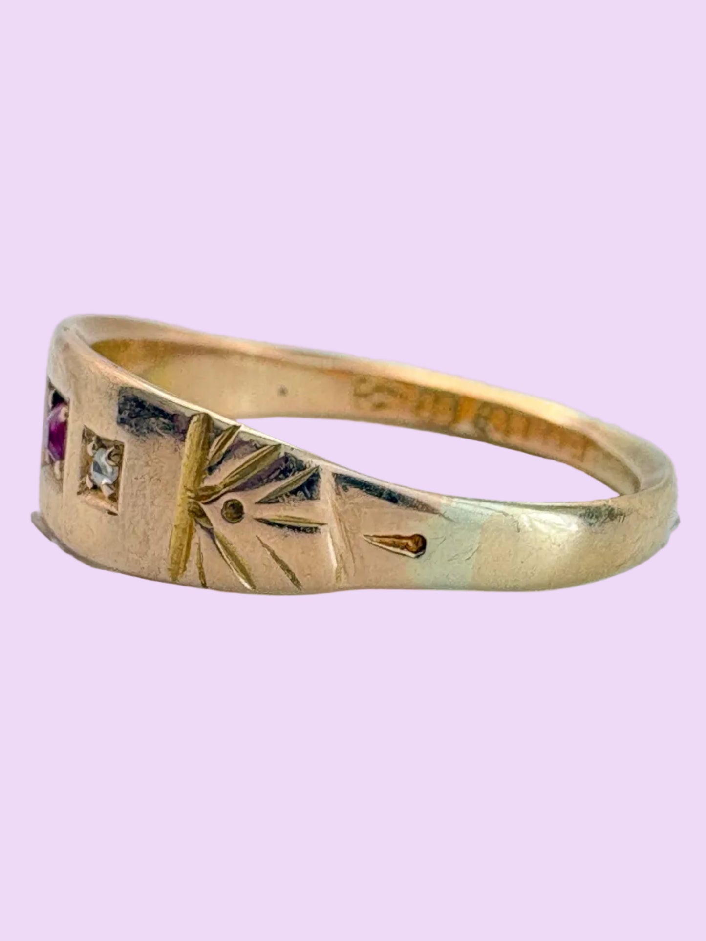 A vintage yellow gold ring with engraved geometric patterns and a small pink gemstone on the left side, shown on a light gray background.