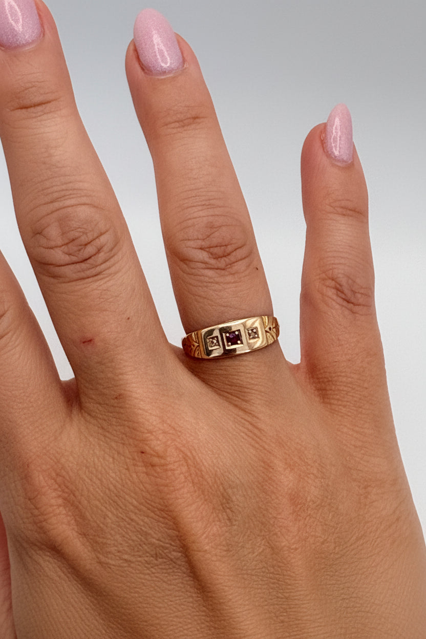 A gold ring with a geometric, block-like pattern and small dark square accents, shown worn on a person's ring finger against a light gray background.