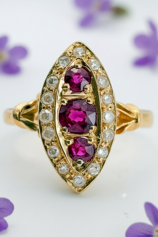 An antique gold ring featuring three oval-cut deep pinkish-purple rubies set in a gold marquise-shaped halo, surrounded by small round diamonds, on a white background.