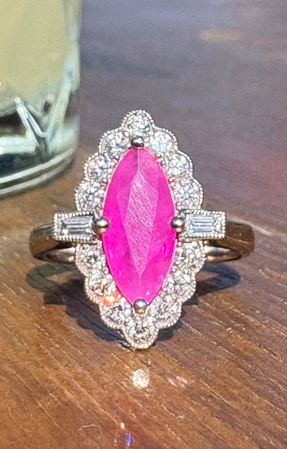A vintage-style ring featuring a large marquise-cut bright pink ruby center stone, surrounded by a scalloped halo of small clear stones, with baguette side stones on a gold-toned band, resting on a wooden surface.