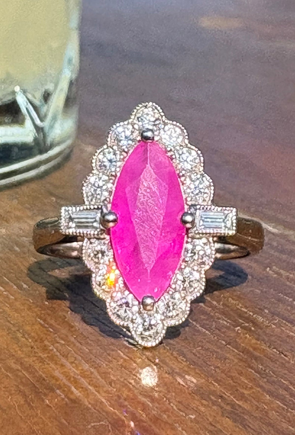 A platinum set ring featuring a prominent marquise-cut bright pink ruby surrounded by diamonds, displayed on a wooden surface with a cocktail in the background.