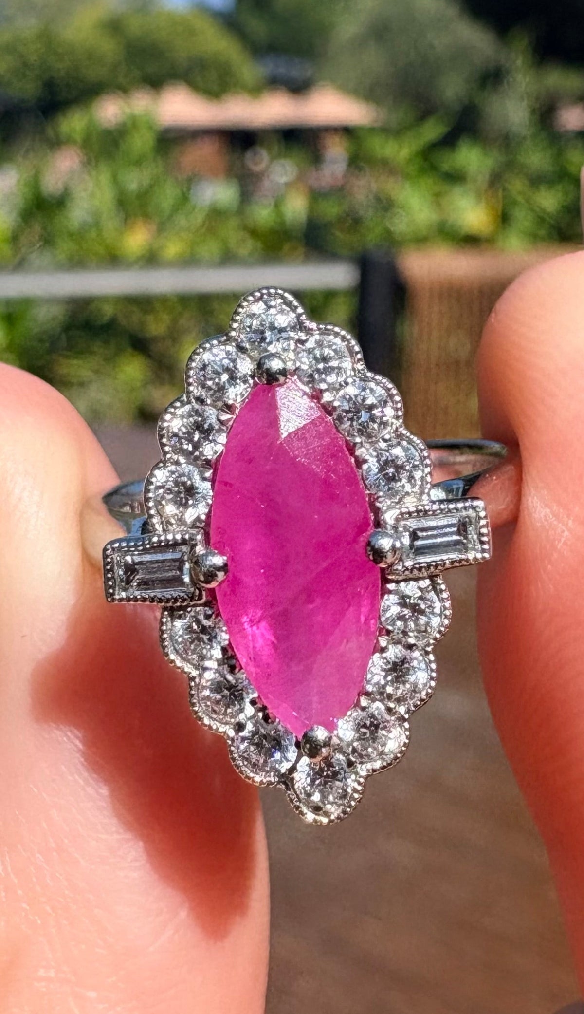 A platinum ring featuring a marquise-cut pink ruby centre stone surrounded by a halo of diamonds, held between two fingers against a blurred outdoor garden background.