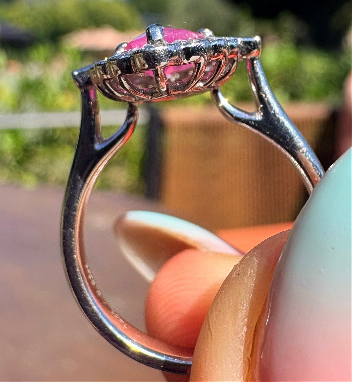 A close-up shot of a platinum ring with a pink ruby and diamond cluster, held between fingers against a blurred outdoor background.