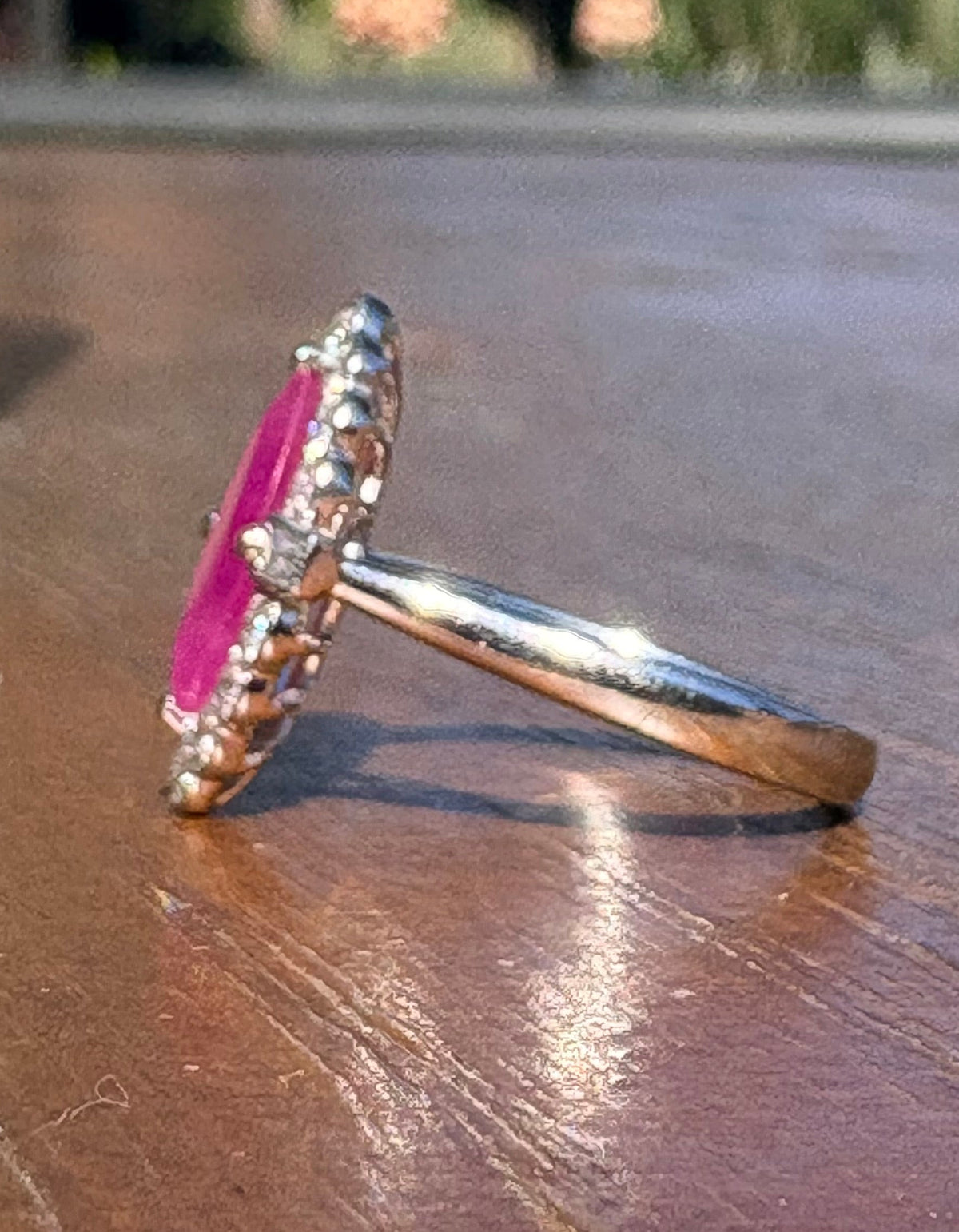 A silver ring with a vibrant pink oval gemstone and small clear accent stones, resting on a wooden table with an outdoor patio background.