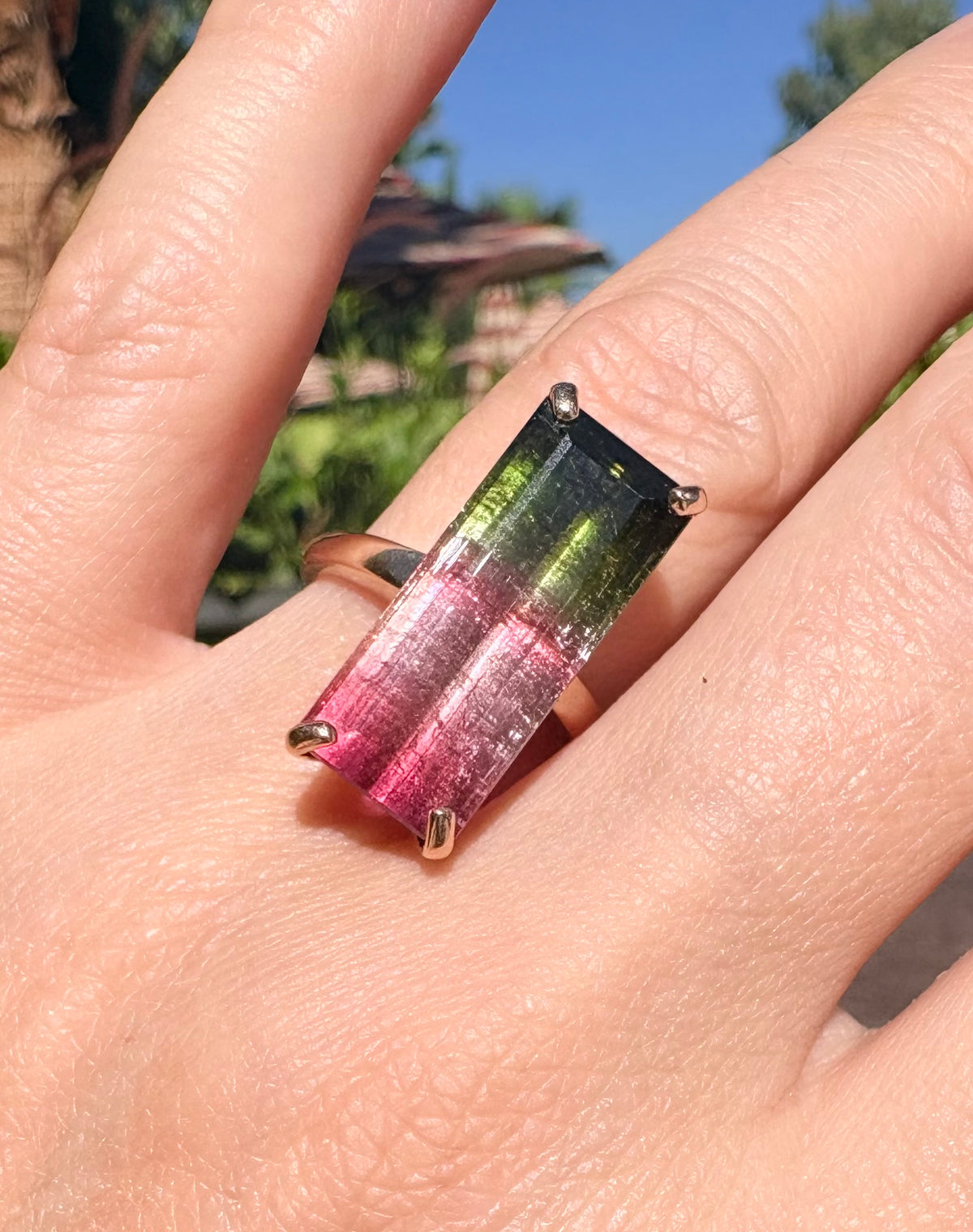 A rectangular gradient gemstone ring with pink, green, and dark blue tones in a gold setting, worn on a person's finger outdoors.