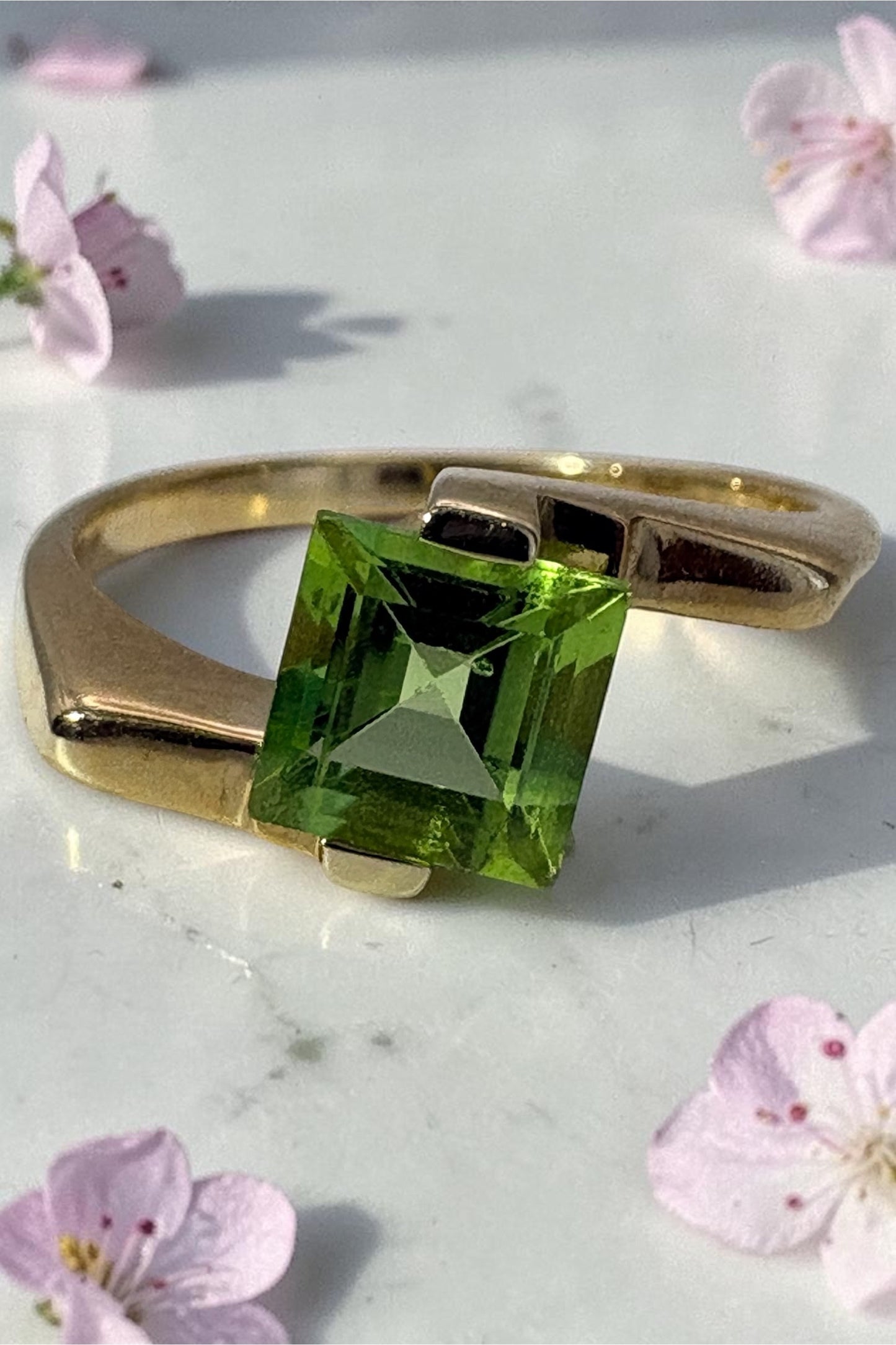 A gold ring featuring a square-cut peridot gemstone, displayed on a light marble surface with scattered pink flowers.