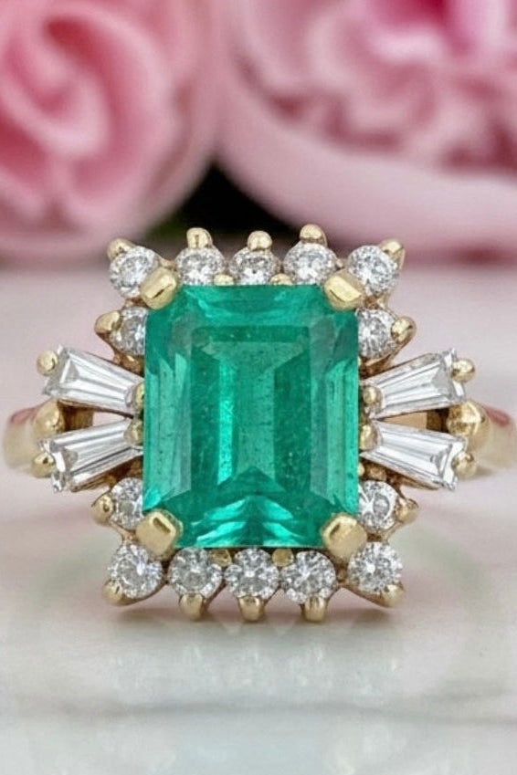 An emerald-cut green gemstone ring with a gold-tone halo and baguette side stones, displayed on a light marble surface with soft pink flowers blurred in the background.