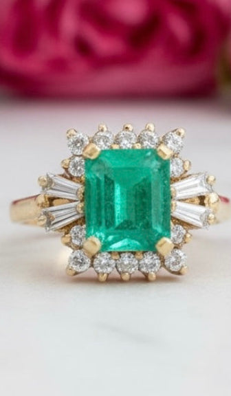 A yellow gold ring featuring a large emerald-cut green gemstone surrounded by small clear stones and baguette accents, photographed on a light surface with a blurred pink rose in the background.