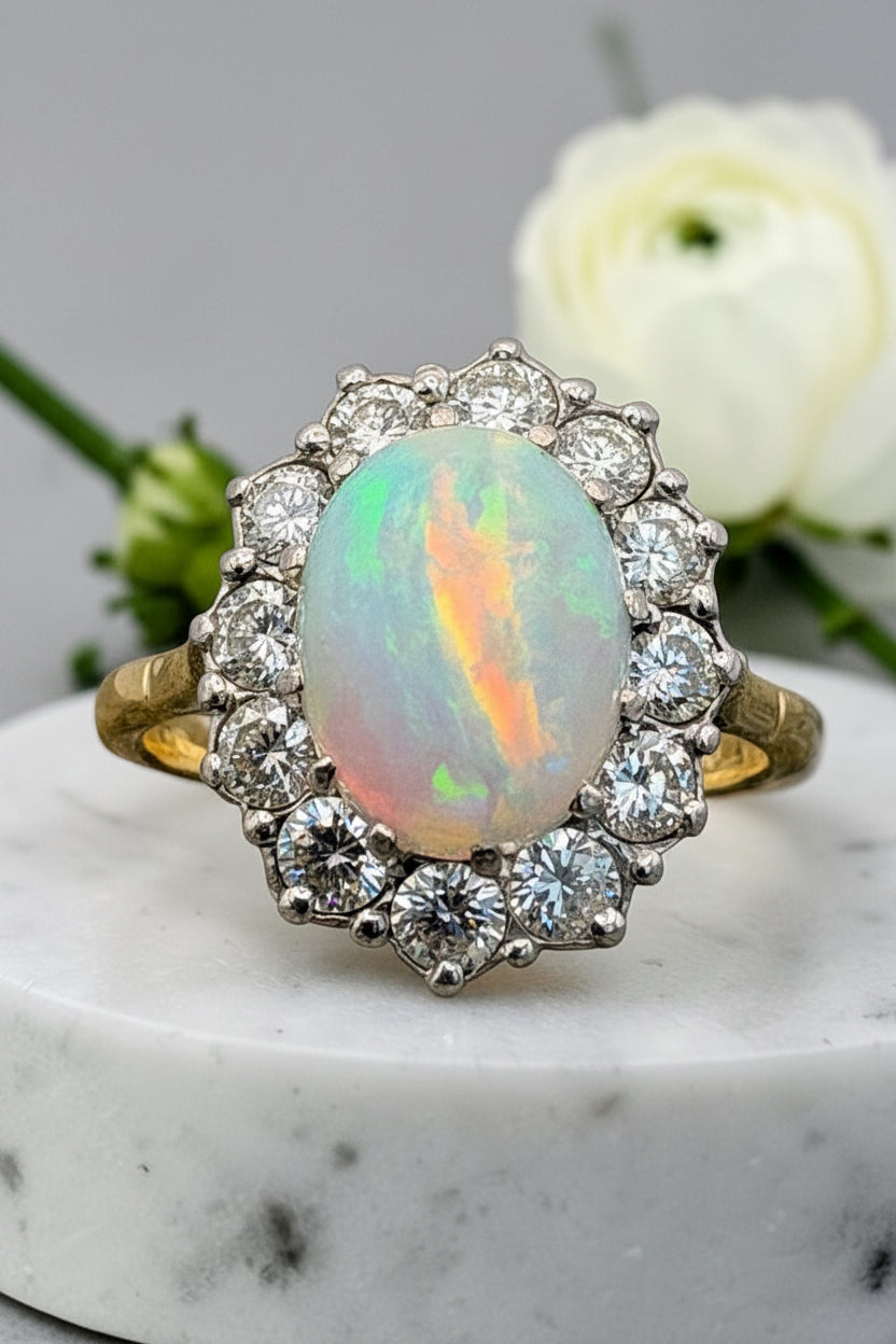 A vintage-style cluster ring featuring a central iridescent opal gemstone surrounded by a halo of diamonds, set on a yellow gold band, displayed on a white marble surface with a white rose in the background.