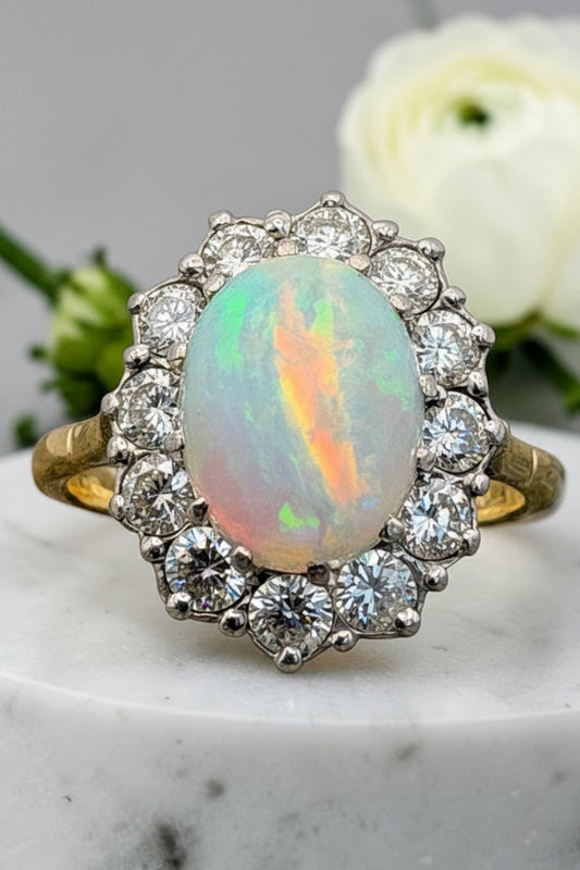 A vintage-style cluster ring featuring a central iridescent opal gemstone surrounded by a halo of diamonds, set on a yellow gold band, displayed on a white marble surface with a white rose in the background.