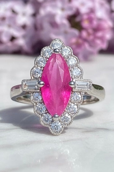 A platinum ring featuring a marquise-cut pink ruby surrounded by a halo of diamonds, displayed on a light marble surface with blurred pink flowers in the background.
