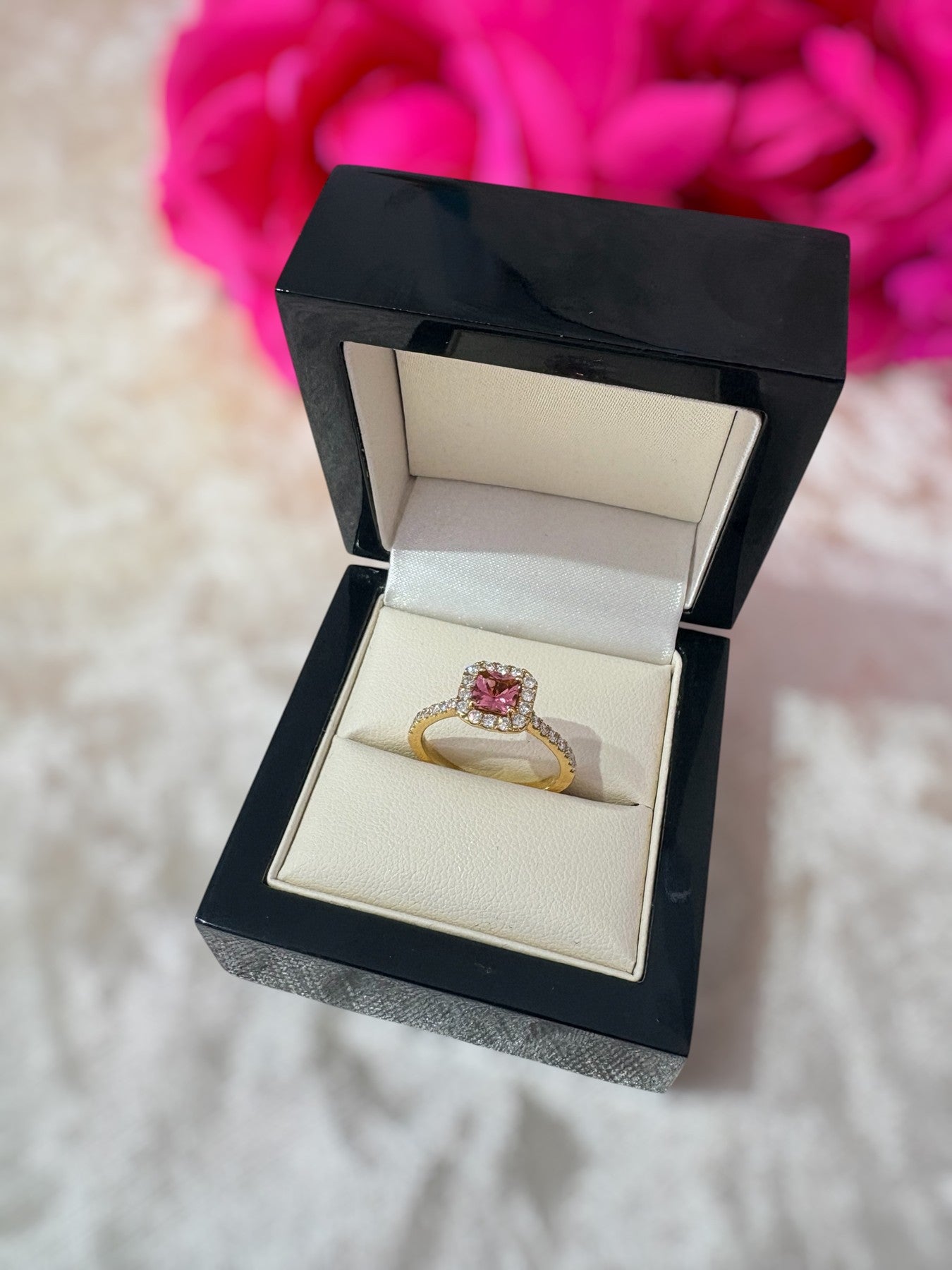 Gold ring with a pink gemstone in a black jewelry box on a soft surface with pink flowers in the background.